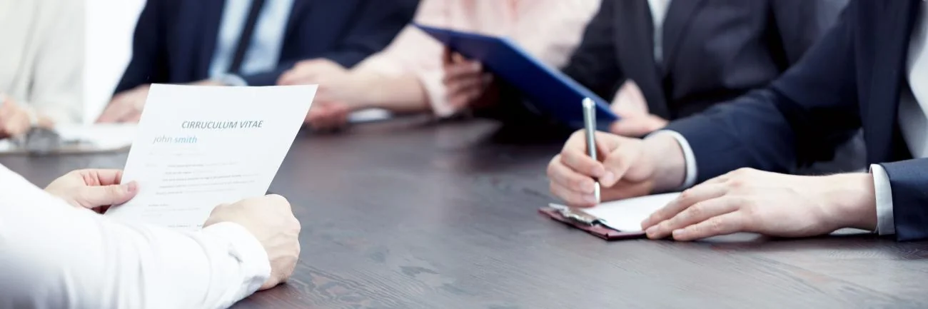 A recruiter reviews a printed CV across the table from a hiring panel